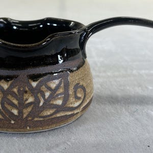 May include: A small, ceramic creamer with a black rim and handle. The body is tan with a brown leaf design. The creamer has a curved handle and a spout for easy pouring. A handmade kitchenware item.