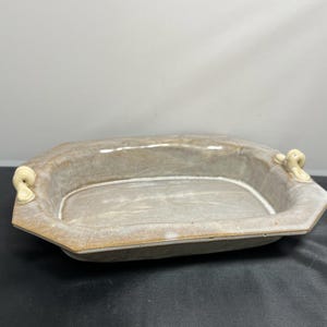 May include: A light brown ceramic serving dish with two small, off-white handles on either side. The dish has a slightly rounded rectangular shape with a slightly raised rim.