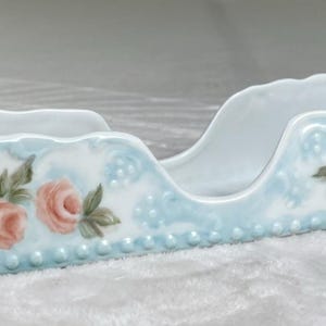 Puede incluir: Un plato de porcelana blanca con un diseño floral azul claro. El plato presenta rosas rosas y hojas verdes, con un borde festoneado y asas. La base tiene un ribete de cuentas.