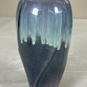 May include: A ceramic vase with a distinctive glaze. The vase transitions from a dark blue base to a lighter blue and white at the top, with a dripped effect. The vase has a smooth, glossy finish and a slightly flared opening.