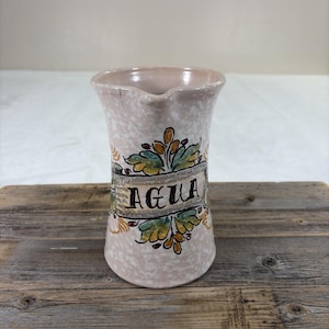 Könnte beinhalten: Ein Keramikkrug mit hellrosa gesprenkelter Oberfläche und ausgestelltem Rand. Die Vorderseite zeigt ein handgemaltes Banner mit dem Wort AGUA in schwarzer Schrift, umgeben von dekorativen grünen und gelben Blattmotiven oben und unten.