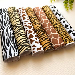 May include: Seven cylindrical rolls of fabric featuring various animal print patterns, including zebra stripes, tiger stripes, cow spots, giraffe spots, and leopard spots, arranged side by side in a row on a light-colored surface.