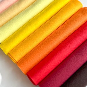 May include: An angled display of rolled felt sheets in a variety of colours. The colours include peach, yellow, orange, red, burgundy, and brown. The felt has a soft texture, ideal for craft projects and creative endeavours.