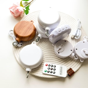 Op de afbeelding: Een verzameling ronde motoren voor babymobielen in witte en houtnerfafwerkingen, voorzien van ophanghaken, bedieningsknoppen en een witte afstandsbediening met het label HLEEDUO. Inclusief een witte USB-oplaadkabel en een klein bruin stoffen label.