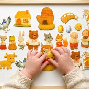 Pode incluir: Uma coleção de autocolantes de feltro com uma casa, uma luva, uma ponte, um sol, ovos e vários animais e pessoas com roupas tradicionais. As figuras incluem um urso, raposa, lobo, coelho, sapo, rato, vaca e duas pessoas idosas.