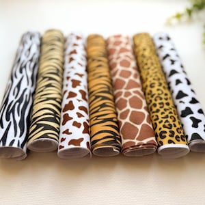 May include: Seven rolls of fabric arranged side by side, each featuring a distinct animal print pattern including zebra stripes, tiger stripes, giraffe spots, leopard spots, and cow hide patterns in black, white, brown, and golden yellow tones.