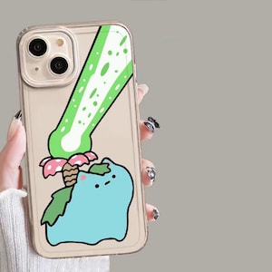 May include: Clear phone case featuring a cartoon illustration of a blue creature with green leaves and a pink and white mushroom. A green and white design is above the creature. The case has a gold-colored border.