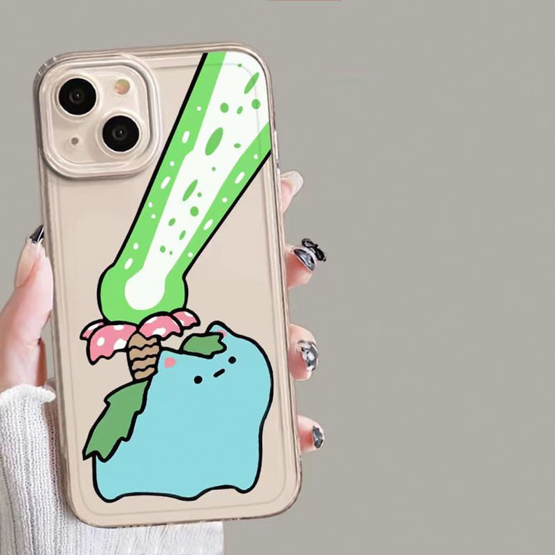 May include: Clear phone case featuring a cartoon illustration of a blue creature with green leaves and a pink and white mushroom. A green and white design is above the creature. The case has a gold-colored border.