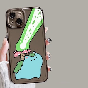 May include: A clear phone case with a cartoon illustration of a blue creature with green leaves, a pink and white mushroom, and a green plant. The case has a brown tint and a black border.