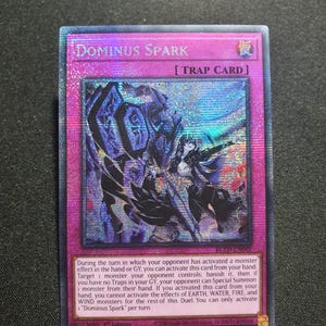 Puede incluir: Una carta trampa holográfica Dominus Spark del juego de cartas coleccionables Yu-Gi-Oh!. La carta presenta un borde magenta, una ilustración central de un personaje de pelo oscuro con un gran artefacto azul y texto detallado del efecto.