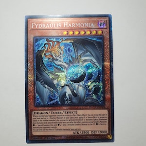 Puede incluir: Carta coleccionable holográfica con "Fydraulis Harmonia". La carta tiene un diseño azul y plateado con una ilustración de criatura parecida a un dragón. Texto incluye tipo de carta, efectos y estadísticas: ATK/2500 DEF/2000.