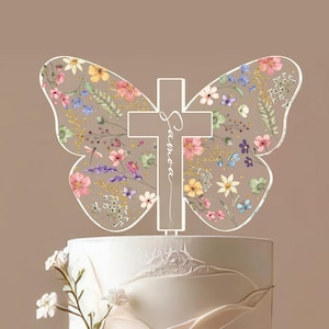 May include: A clear acrylic cake topper in the shape of a butterfly with a cross in the center. The butterfly wings are decorated with colorful floral patterns. The cross has the name "Samora" written on it.