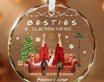 May include: A circular glass ornament with a white ribbon, featuring a graphic of two people in red plaid pajamas lying on a brown sofa. The text reads B.E.S.T.I.E.S. I'LL BE THERE FOR YOU, with the names ROXANA and CASEY printed below the sofa.