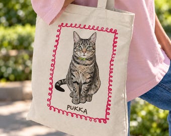 May include: A beige canvas tote bag featuring a centered illustration of a brown tabby cat wearing a small green collar. The cat is framed by a red, spiraled rectangular border, with the name PUKKA printed in black capital letters below.