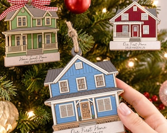 May include: Three house-shaped Christmas ornaments featuring a green, red, and blue house. Each ornament has a white base with printed text: Home Sweet Home 2392 Sandstone, Our First Home 1618 Hazen Ln, and Our First Home 148 Wilson St.