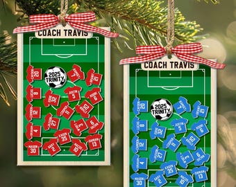 Könnte beinhalten: Zwei rechteckige Holzanhänger mit einer grünen Fußballfeldgrafik, dem Text COACH TRAVIS und einem Fußball mit 2025 TRINITY. Ein Anhänger zeigt rote Trikots mit Namen und Nummern, der andere blaue Trikots, beide mit roten Vichy-Schleifen verziert.