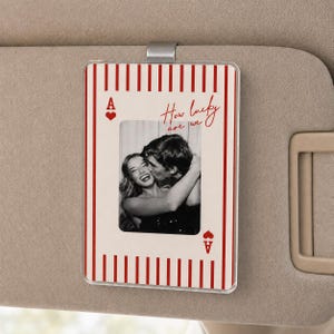 May include: A rectangular car visor clip featuring a playing card design with red and white vertical stripes. The card displays the letter A and a heart symbol, with the text How lucky are we and a black and white photograph of a couple.