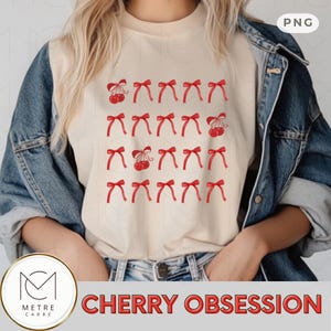 Pode incluir: Uma camiseta creme com um padrão de laços e cerejas vermelhas. O texto "CHERRY OBSESSION" é exibido em letras vermelhas na parte inferior. A modelo está usando uma jaqueta jeans.