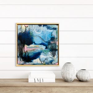 May include: A square abstract painting in a gold frame featuring layered shades of navy blue, light blue, and white with accents of pink and green. The composition includes expressive brushstrokes, dark charcoal lines, and organic shapes.