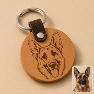 May include: A round tan leather keychain featuring a dark brown leather strap with a silver metal snap and a silver split ring. The leather surface is laser-engraved with a detailed line art illustration of a German Shepherd dog face.