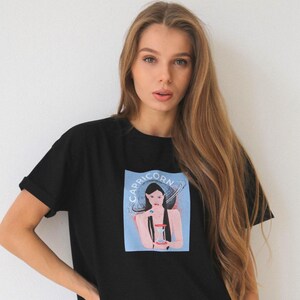 Peut inclure: T-shirt noir avec un imprim&eacute; graphique d'une femme sur fond bleu et le texte "Capricorn" au-dessus d'elle. La femme tient un sablier et est entour&eacute;e d'un anneau de plan&egrave;tes.