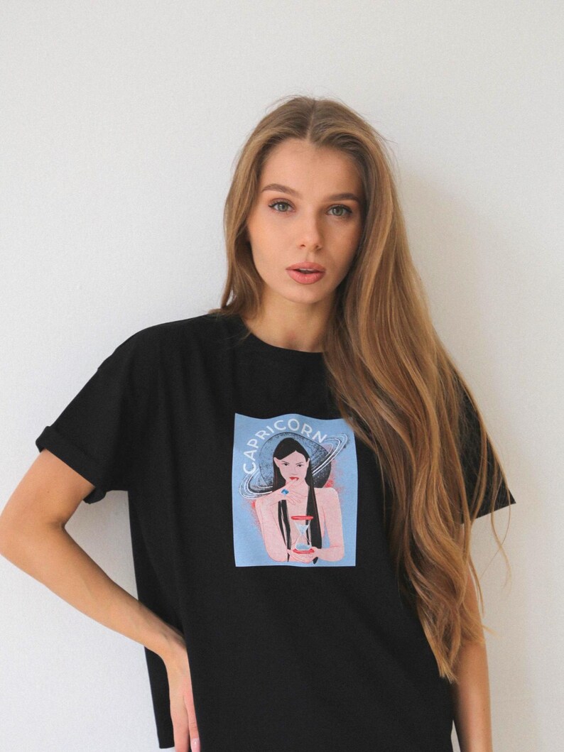 Peut inclure: T-shirt noir avec un imprim&eacute; graphique d'une femme sur fond bleu et le texte "Capricorn" au-dessus d'elle. La femme tient un sablier et est entour&eacute;e d'un anneau de plan&egrave;tes.