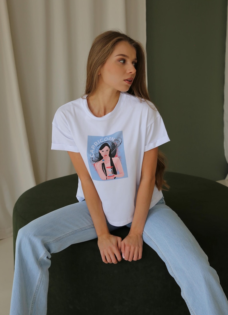 Peut inclure: Un t-shirt blanc avec un imprim&eacute; repr&eacute;sentant une femme avec le mot "Capricorne" imprim&eacute; dessus. La femme porte une robe bleue et une couronne. Le t-shirt est port&eacute; par une personne assise sur une chaise verte.