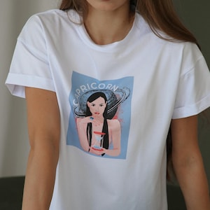 Peut inclure: T-shirt blanc avec un graphique bleu d'une femme aux cheveux fonc&eacute;s et une bague bleue au doigt. Le graphique comprend &eacute;galement le mot "Capricorne" et une plan&egrave;te annulaire.
