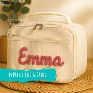 May include: Cream-colored lunch bag with a vertical ribbed texture, a top handle, and a front pocket. The name "Emma" is embroidered in pink cursive letters. The bag has a zipper closure and is sitting on a woven mat.
