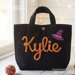 Op de afbeelding: Een zwarte corduroy tas met de naam Kylie in oranje garenletters op de voorzijde. De tas is voorzien van een goudkleurige drukknoopsluiting aan de bovenzijde en een paarse glitterende heksenhoedapplicatie rechts van de naam.