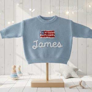 May include: A light blue knit sweater with the name "James" embroidered in white. The sweater features a small, colorful patch resembling a flag. The sweater is displayed on a wooden stand, suggesting it's a children's garment.