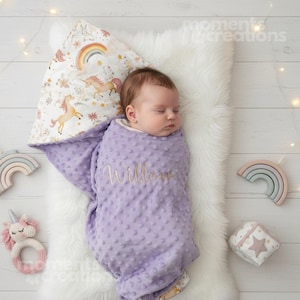 May include: A baby sleeping in a lavender dimpled swaddle blanket embroidered with the name "Willow." The blanket rests on a white faux fur rug, with a unicorn-themed pillow and wooden toys.