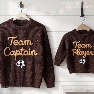 May include: Two brown knit sweaters with the words "Team Captain" and "Team Player" in yellow script. Each sweater has a soccer ball patch. The sweaters are hanging on wooden hangers against a white wall.