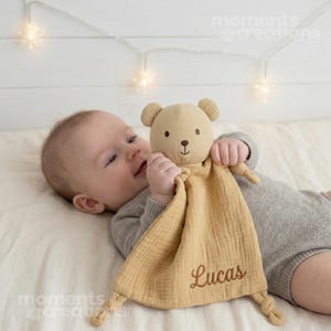 May include: A tan baby comforter featuring a teddy bear head and the embroidered name "Lucas". The bear has small ears, a brown nose, and black eyes. The blanket is made of a soft, textured fabric.