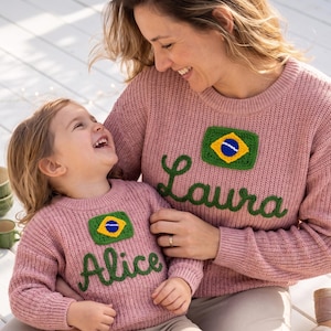 May include: Two matching pink sweaters, one worn by an adult and the other by a child. Each sweater features a crochet patch of the Brazilian flag and a name in green script: "Laura" and "Alice".