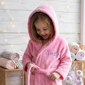May include: A soft, plush, bright pink hooded bathrobe featuring long sleeves, a matching tie belt, and the name Emily embroidered in purple cursive script on the left chest area. The robe is made of a fuzzy, textured fabric with a hood.