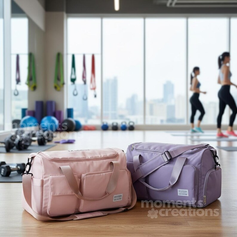 May include: Two duffel bags in a gym setting. One is light pink, the other is lavender. Both have shoulder straps and multiple pockets. The bags are on a wooden floor, with exercise equipment and people in the background.