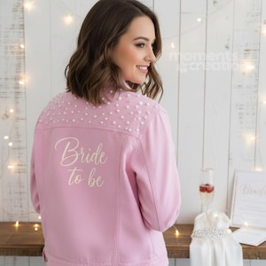 K&ouml;nnte beinhalten: Hellrosa Jeansjacke mit Perlenbesatz auf den Schultern und dem Schriftzug "Bride to be" in Schreibschrift. Die Jacke ist Teil eines Brautthemas, mit einem Sektglas und einem Brautkleid auf einer Holzoberfl&auml;che.