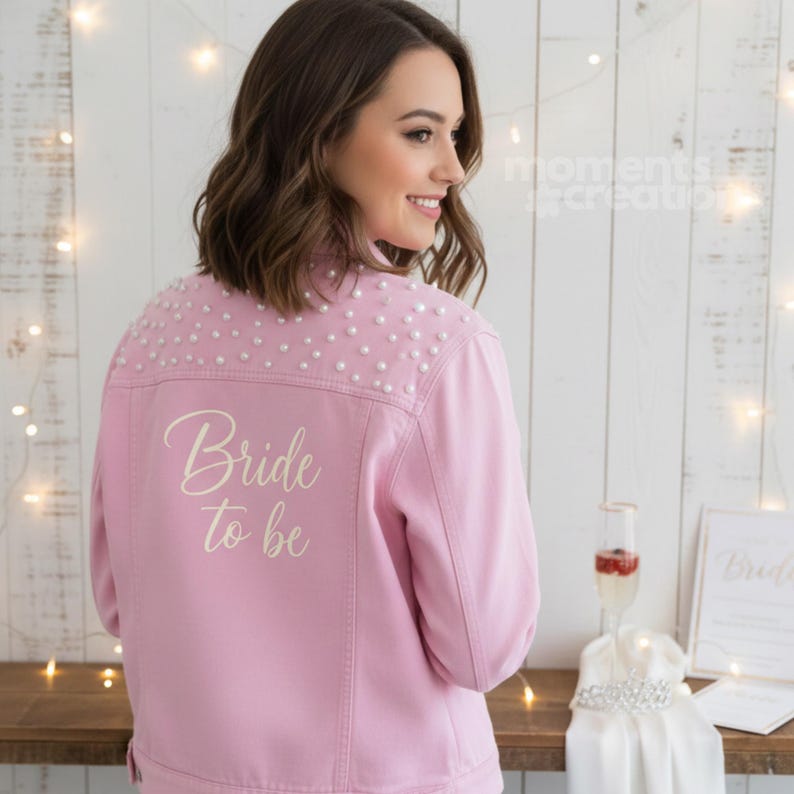 K&ouml;nnte beinhalten: Hellrosa Jeansjacke mit Perlenbesatz auf den Schultern und dem Schriftzug "Bride to be" in Schreibschrift. Die Jacke ist Teil eines Brautthemas, mit einem Sektglas und einem Brautkleid auf einer Holzoberfl&auml;che.