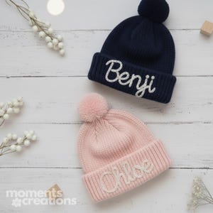 Op de afbeelding: Een marineblauwe gebreide muts met een pompon en de naam Benji in wit geborduurd, en een lichtroze gebreide muts met een pompon en de naam Chloe in wit geborduurd, beide met een geribbelde boord.