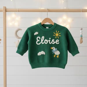 May include: Dark green knitted sweater with the name "Eloise" embroidered in white. The sweater features a sun, clouds, and a duckling design. The sweater is displayed on a wooden hanger.