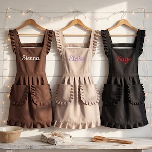 May include: Three aprons hanging on wooden hangers, each featuring ruffled shoulder straps, a ruffled hem, and two ruffled pockets. The brown apron is embroidered with the name Sienna, the beige apron with Eloise, and the black apron with Sage.