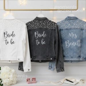 K&ouml;nnte beinhalten: Drei Jeansjacken h&auml;ngen an einer Stange. Eine wei&szlig;e Jacke mit dem Text "Bride to be" befindet sich links. Eine dunkelgraue Jacke mit dem gleichen Text befindet sich in der Mitte. Eine hellblaue Jacke mit "Mrs. Bennett" befindet sich rechts. Alle Jacken haben dekorative Verzierungen.