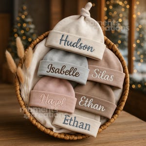 May include: A selection of personalised baby hats in a wicker basket. The hats are in various pastel shades, each embroidered with a name in a matching colour. Names include Hudson, Isabelle, Silas, Hazel, and Ethan.