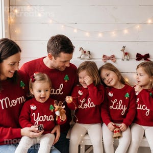 Könnte beinhalten: Eine Gruppe von Personen in passenden roten Strickpullovern mit weiß gestickten Namen wie Mom, Yara, Lola, Lily und Lucy. Die Pullover sind mit kleinen Applikationen in Form von Weihnachtsmann-Gesichtern und grünen Tannenbäumen verziert.
