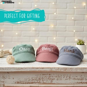 May include: Three corduroy caps in green, pink, and blue, each embroidered with a name: "Ozzy", "Benji", and "Hank". The caps are displayed on a white wooden surface. The text "PERFECT FOR GIFTING" is above the caps.