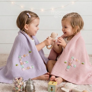 Op de afbeelding: Twee jonge kinderen gehuld in zachte dekens, één lavendelkleurig en één roze, geborduurd met bloemen- en bijenontwerpen. De dekens zijn over de schouders van de kinderen gedrapeerd. Eén kind houdt een teddybeer-speeltje vast. Een pot met bloemen en een houten blok staan op de voorgrond.