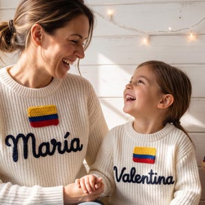 Pode incluir: Camisolas creme a condizer com patches da bandeira colombiana e os nomes "Maria" e "Valentina" bordados em azul marinho. Uma mãe e sua filha usam as camisolas e sorriem uma para a outra.