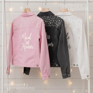 K&ouml;nnte beinhalten: Drei Jeansjacken in Rosa, Anthrazit und Wei&szlig; h&auml;ngen an einem wei&szlig;en Gestell. Die rosa Jacke hat den Text "Maid of Honor" und Perlenverzierungen. Die anthrazitfarbene Jacke hat Perlenverzierungen und den Text "Mrs. Super". Die wei&szlig;e Jacke hat Perlenverzierungen und den Text "Bride Forever".