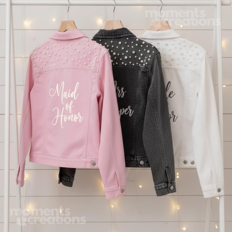 K&ouml;nnte beinhalten: Drei Jeansjacken in Rosa, Anthrazit und Wei&szlig; h&auml;ngen an einem wei&szlig;en Gestell. Die rosa Jacke hat den Text "Maid of Honor" und Perlenverzierungen. Die anthrazitfarbene Jacke hat Perlenverzierungen und den Text "Mrs. Super". Die wei&szlig;e Jacke hat Perlenverzierungen und den Text "Bride Forever".
